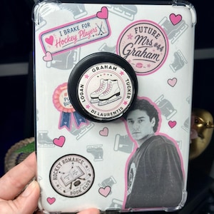 Könnte beinhalten: Eine transparente Handyhülle mit Hockey-Aufklebern, darunter ein rosa 'I BRAKE FOR Hockey Players'-Schild, ein 'FUTURE Mrs. Graham #44'-Abzeichen und ein schwarzer, runder Handyhalter mit Schlittschuhen sowie den Namen Logan und Tucker.
