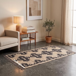May include: A rectangular area rug featuring a repeating dark charcoal geometric pattern on a natural beige background, finished with short fringe on both ends. The rug displays the text JuteRugHouse in the bottom left corner.