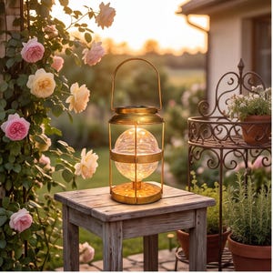 May include: A gold-toned metal lantern with a spherical glass center containing warm glowing lights. The lantern features a wire frame, a top handle, and a circular base, sitting on a wooden stool in an outdoor garden setting with roses.