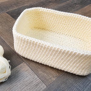 Könnte beinhalten: Ein rechteckiger, cremeweißer Häkelkorb mit dichtem, strukturiertem Maschenbild und abgerundeten Ecken, begleitet von drei passenden Knäueln cremeweißem Garn, die neben dem Behälter auf einer Oberfläche in dunkler Holzoptik liegen.