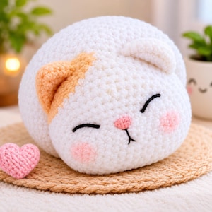 Könnte beinhalten: Ein rundes, weißes, gehäkeltes Katzen-Plüschtier mit einem orangefarbenen Ohr, geschlossenen Augen, einer kleinen rosa Nase und rosa Wangen. Ein kleines, separates rosa gehäkeltes Herz liegt links neben der Katze auf einer runden, beigen Matte.