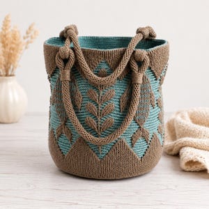 Könnte beinhalten: Eine Beuteltasche aus gewebtem Garn mit einem zweifarbigen Muster in Braun und Blaugrün. Das Design zeigt ein sich wiederholendes Blattmotiv und geometrische Formen. Sie verfügt über zwei dicke, geflochtene Griffe am oberen Rand.