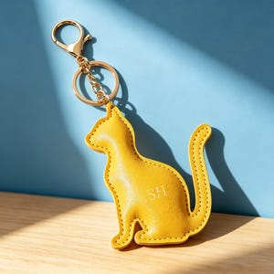 May include: A bright yellow cat-shaped keyring made of a textured material with visible stitching along the edges. The keyring features the letters SH embossed in the centre and is attached to a gold-toned metal split ring and a lobster clasp.