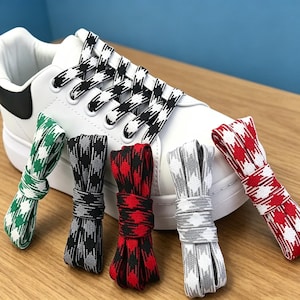 Könnte beinhalten: Ein weißer Sneaker mit schwarz-weißem Hahnentrittmuster in den Schnürsenkeln. Vier weitere Paare flacher Schnürsenkel sind davor abgebildet, mit grünen, schwarzen, roten und grauen Hahnentrittmustern auf weißem Grund.