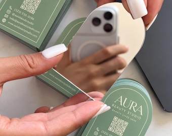 May include: Arch-shaped business cards with a sage green background and white text reading AURA BEAUTY STUDIO. The cards feature a QR code, phone number, social media handle, email, and website. One card is reflective, showing a smartphone reflection.