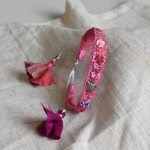 Puede incluir: Una pulsera de tela rosa con intrincados bordados florales multicolores a lo largo de la banda. La pulsera incluye dos borlas de tela colgantes en tonos rosa claro y magenta intenso, unidas por cordones blancos finos a la correa principal.