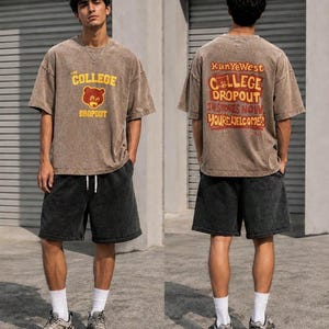 May include: A brown acid-wash t-shirt featuring a bear graphic and the text THE COLLEGE DROPOUT on the front, and the text KANYE WEST COLLEGE DROPOUT IN STORES NOW YOU'RE WELCOME on the back, paired with black acid-wash shorts and white socks.