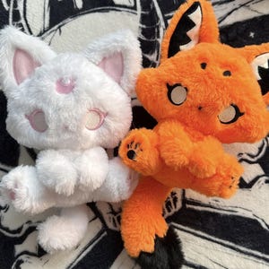Könnte beinhalten: Zwei Plüschtiere, eine weiße Katze und ein orangefarbener Fuchs. Die weiße Katze hat rosa gestickte Augen und eine Mondsichel auf der Stirn. Der orangefarbene Fuchs hat schwarz gespitzte Ohren, schwarze Pfotenballen und einen schwarz gespitzten Schwanz.