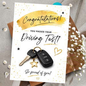 Op de afbeelding: Een witte wenskaart met gouden glitteraccenten en de tekst Congratulations! YOU PASSED YOUR DRIVING TEST! So proud of you! De kaart toont een zwarte autosleutel en een metalen sleutel aan een goudkleurige sleutelhanger in de vorm van een ster.
