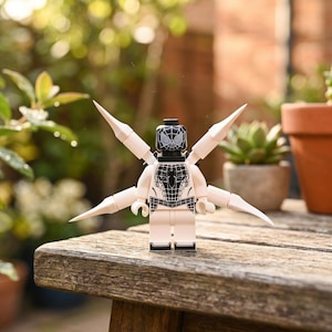 Könnte beinhalten: Eine weiß-schwarze Kunststoff-Minifigur mit Spinnennetz-Muster auf Torso und Kopf sowie vier spitzen weißen Gliedmaßen, die vom Rücken abstehen. Die Figur hat eine schwarze Maske mit weißen Augenlinsen und ein detailliertes Spinnenemblem auf der Brust.