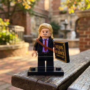 Könnte beinhalten: Eine Lego-Minifigur mit blondem Haar, bekleidet mit einem schwarzen Sakko über einem violetten Hemd und einer roten Krawatte. Ein brauner Riemen verläuft über die Brust. Die Figur hält ein schwarzes Schild mit der goldenen Aufschrift NELSON AND MURDOCK.