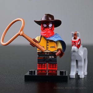 Peut inclure: Une figurine de bloc de construction en plastique habillée en cow-boy avec un masque rouge, un chapeau marron, un gilet jaune et une cape bleue, tenant un lasso marron. À côté se trouve un petit cheval gris avec un masque à motif toile d'araignée.