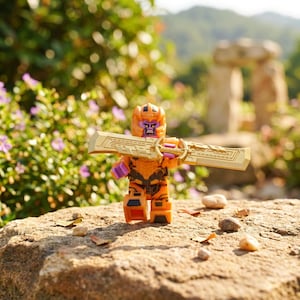 Könnte beinhalten: Eine kleine orange-schwarze Kunststoff-Actionfigur mit violettem Gesicht, die einen Helm trägt und ein großes, breites, goldfarbenes Schwert mit komplizierten mechanischen Details auf der Klinge und einem runden Parier in der Mitte hält.