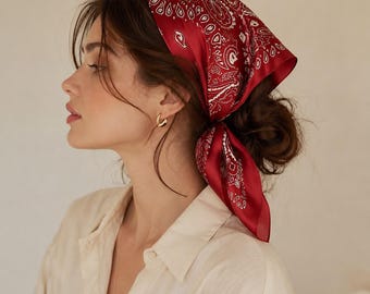 May include: A square head scarf featuring a deep red base color with a white paisley pattern. The fabric has a smooth, satin-like finish and is tied at the back of the head in a triangular fold, with the ends hanging down behind the neck.