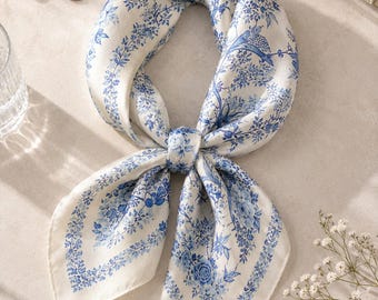May include: A square silk scarf featuring a white base with a detailed blue floral and botanical print. The scarf is tied in a knot at the center, displaying intricate patterns of flowers, leaves, and branches arranged in a symmetrical design.