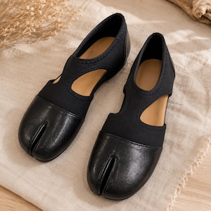 May include: A pair of black split-toe shoes featuring a smooth leather-like front section and a flexible fabric upper. The design includes a distinctive cutout detail along the sides and a tan interior lining, showcasing a low-profile construction.