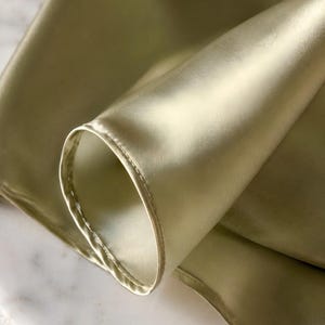 May include: A smooth, light olive green fabric with a subtle sheen and a soft, folded texture. The material features a finished, stitched hem along the curved edge, showcasing the reflective quality of the textile and its fluid, draping nature.