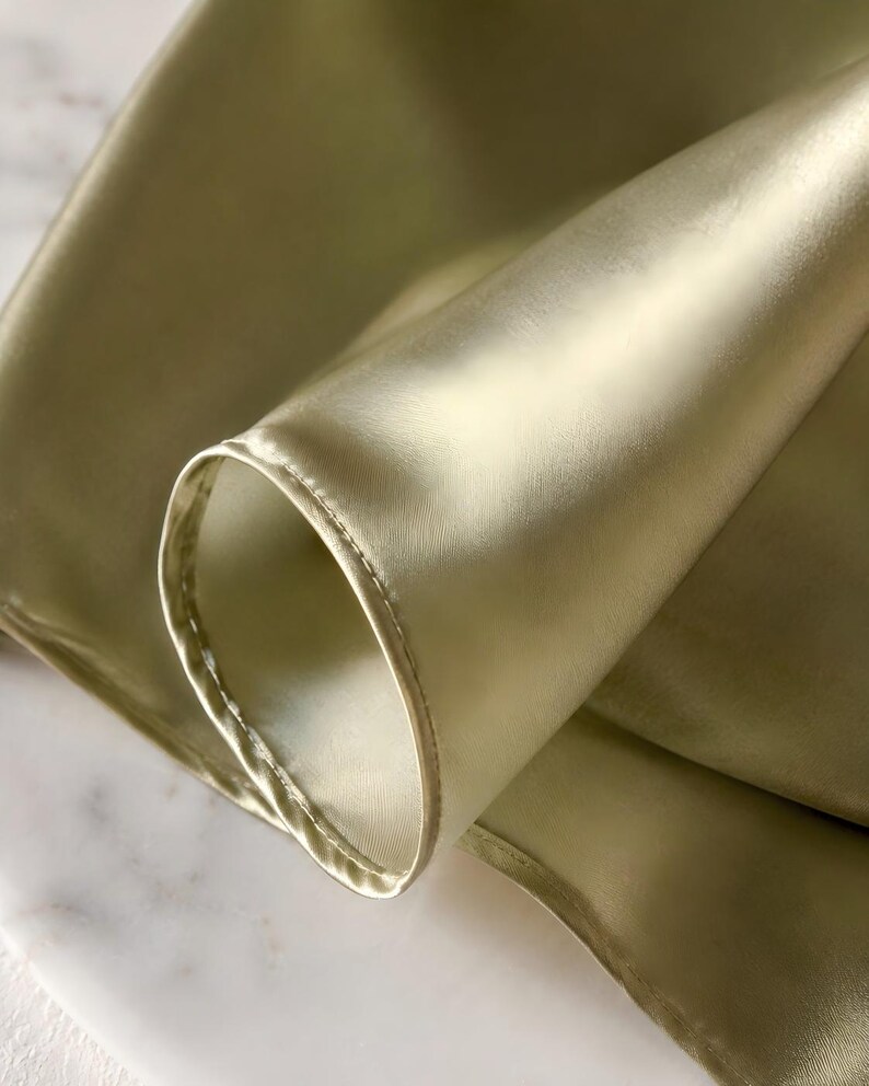 May include: A smooth, light olive green fabric with a subtle sheen and a soft, folded texture. The material features a finished, stitched hem along the curved edge, showcasing the reflective quality of the textile and its fluid, draping nature.