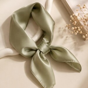 May include: A sage green square scarf with a smooth, lustrous finish, tied in a simple knot. The fabric has a soft sheen and a fluid drape, creating gentle folds and shadows throughout the material, showcasing a solid, muted green color.