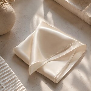 May include: A square, off-white satin pocket square or scarf with a smooth, reflective surface and finished stitched edges. The fabric is folded into a triangular shape, showcasing its soft sheen and light-catching texture in a neutral tone.