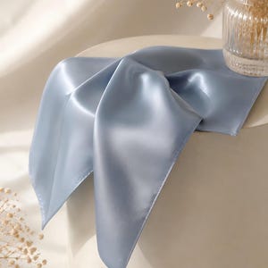 May include: A square scarf in a solid dusty blue color with a smooth, satin-like finish. The fabric is draped over a cylindrical surface, showing its reflective sheen and soft folds. The edges of the scarf are finished with a clean, straight hem.