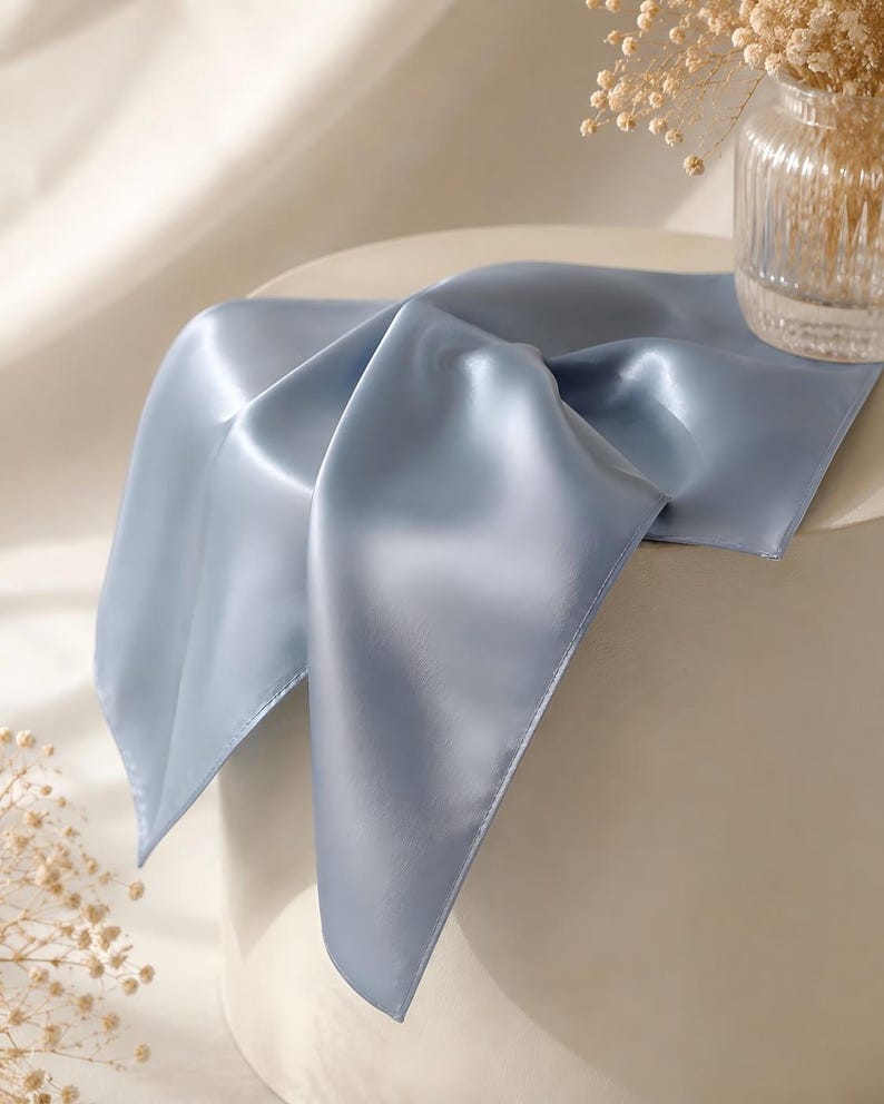 May include: A square scarf in a solid dusty blue color with a smooth, satin-like finish. The fabric is draped over a cylindrical surface, showing its reflective sheen and soft folds. The edges of the scarf are finished with a clean, straight hem.