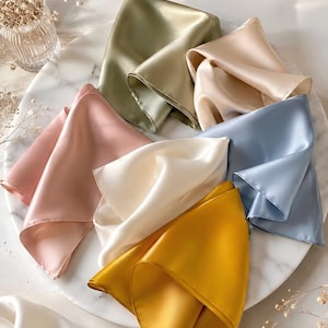 May include: A collection of six square satin scarves in various solid colors including sage green, pale pink, cream, mustard yellow, light blue, and beige. Each scarf features a smooth, lustrous finish and is folded to display its soft, draped texture.
