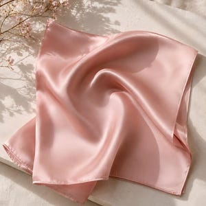 May include: A square-shaped scarf in a solid light pink color with a smooth, lustrous finish. The fabric is folded and draped to create soft, flowing curves and shadows across its surface, highlighting the reflective quality of the material.