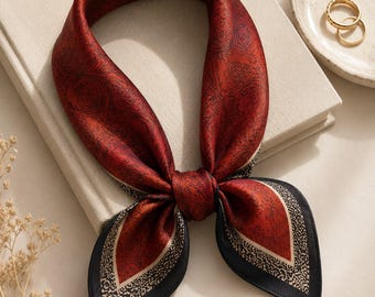 May include: A square scarf featuring a deep red center with a dark paisley pattern, bordered by a cream-colored section with a black speckled design and a solid black edge. The scarf is tied in a knot, displaying a smooth, satin-like texture.