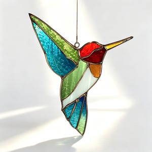 May include: A stained glass hummingbird suncatcher featuring vibrant panels of red, orange, green, blue, and white glass held together by dark metal solder lines. The bird is depicted in flight with wings extended and a long, pointed beak.