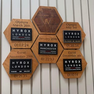 May include: A collection of six hexagonal wooden plaques arranged in a cluster. Each plaque features engraved event details, including locations like Olympia and Excel, dates, and attached rectangular patches with Hyrox London or Manchester branding.
