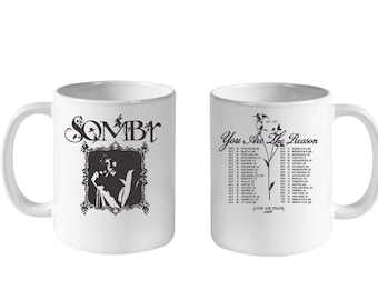 May include: Two white ceramic mugs featuring black graphics. One mug displays the text SOMBY above a framed portrait. The other mug features the text You Are The Reason, a floral illustration, and a list of tour dates and locations.