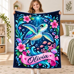 May include: A dark blue fleece blanket featuring a central sea turtle surrounded by vibrant pink hibiscus flowers, colourful coral, small fish, and bubbles. A pink ribbon banner at the bottom displays the name Olivia in black script.