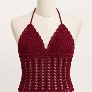 May include: A deep burgundy crochet halter top featuring a triangular bodice with a scalloped edge and a lower section crafted in an openwork mesh pattern. The garment includes thin neck straps and a straight hemline with a matching scalloped finish.