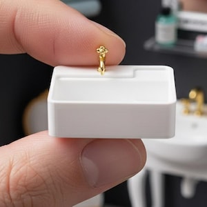 May include: A miniature white bathroom sink basin with a small gold-colored faucet fixture. The rectangular sink features a smooth, matte finish and is held between fingers to demonstrate its small scale relative to a human hand.