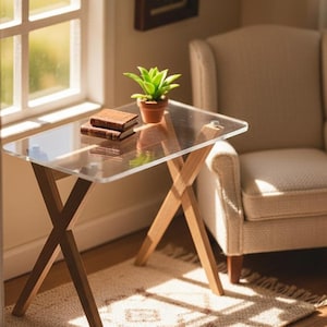 May include: A miniature dollhouse desk featuring a clear acrylic rectangular top supported by light-toned wooden legs in an X-frame design. On the desk sits a small potted green succulent and two stacked miniature books with brown covers.