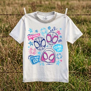 Peut inclure: Un t-shirt à manches courtes blanc cassé avec un imprimé graphique composé d'illustrations d'araignées bleues et roses, incluant des masques, des icônes d'araignées et des étoiles. Le texte "AMAZING", "TEAM UP!" et "GO-WEBS-GO!" est présent.