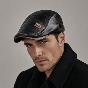 May include: A black flat cap constructed from segmented panels with a smooth finish. The cap features a brown rectangular patch on the side with a metallic emblem and a brown brim underside. An adjustable brown strap with a metal buckle is on the side.