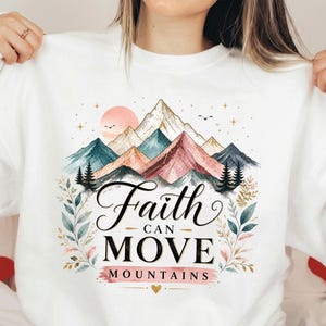 May include: A white sweatshirt featuring a graphic of colorful mountains, pine trees, and a sun. The text reads Faith CAN MOVE MOUNTAINS in black and pink lettering. The design is framed by delicate botanical illustrations of leaves and branches.