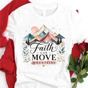 May include: A white t-shirt featuring a graphic of colorful mountains, pine trees, and a sun. The text "Faith CAN MOVE MOUNTAINS" is printed in black and pink lettering in the center, surrounded by delicate botanical illustrations and stars.