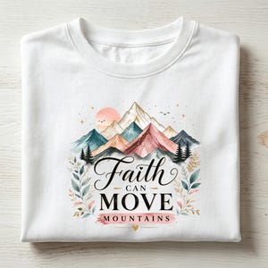 May include: A white t-shirt featuring a graphic print of colorful watercolor mountains, a sun, and pine trees. The text "Faith CAN MOVE MOUNTAINS" is printed in black and pink lettering, surrounded by delicate botanical leaf illustrations.
