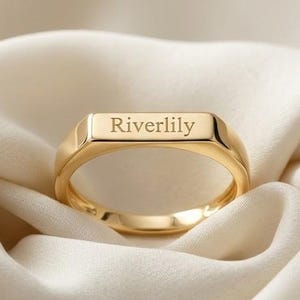 Op de afbeelding: Een gouden zegelring met een plat rechthoekig bovenoppervlak gegraveerd met de tekst Riverlily. De ring heeft een gepolijste metalen afwerking en een gladde band. De tekst Riverlily Jewels wordt weergegeven in het bovenste deel.