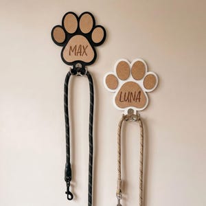 Op de afbeelding: Twee wandhouders voor hondenriemen in de vorm van pootafdrukken. De linkerhouder is zwart met een bruin centrum gegraveerd met de naam MAX. De rechterhouder is wit met een bruin centrum gegraveerd met de naam LUNA. Elk heeft een metalen haak.