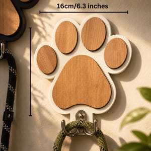 Op de afbeelding: Een wandhaak in de vorm van een pootafdruk met een wit frame en lichtbruine houten inzetstukken. De breedte is 16 cm. Aan de onderkant zit een zilverkleurige metalen dubbele haak met daaraan een groene gevlochten touwlijn.