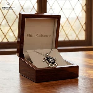 Op de afbeelding: Een zilverkleurige schildpadhanger aan een ketting met een zwarte inleg in het schild, gepresenteerd in een open, gepolijste donkerbruine houten doos. De binnenkant van het deksel bevat de tekst Elite Radiance in donkerbruin op een crèmekleurige achtergrond.