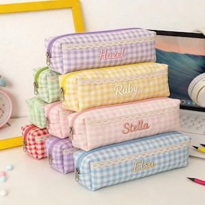 Peut inclure: Une pile de trousses rectangulaires à motifs vichy variés, notamment violet, jaune, rose, bleu, vert et rouge. Chaque trousse présente une bordure en dentelle blanche sur le dessus, une fermeture éclair en métal et un prénom brodé en cursive.