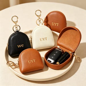 May include: Four semi-circular zippered pouches with gold-toned key rings. One black pouch is embossed with 'we', while two tan and one cream pouch are embossed with 'UYT'. One tan pouch is open, revealing a black car key fob and a small cylinder.