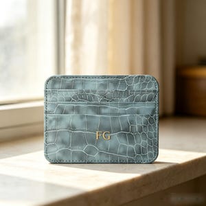 Peut inclure: Un porte-cartes rectangulaire de couleur bleu-gris sourd avec un motif texturé en peau de crocodile. L'avant présente trois fentes horizontales pour cartes et les initiales FG embossées en couleur dorée au centre de la partie inférieure.