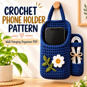 Pode incluir: Um organizador de parede de crochê azul-escuro com bolso para celular e suporte lateral para cabos. A frente apresenta uma aplicação de margarida branca com folhas verdes. Texto: CROCHET PHONE HOLDER PATTERN Wall Hanging Organizer PDF.