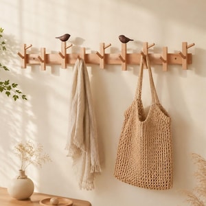 May include: A light wood wall-mounted coat rack featuring multiple pegs and two dark brown bird-shaped decorative accents. A beige linen scarf and a tan woven mesh tote bag are hanging from the pegs of the rack.